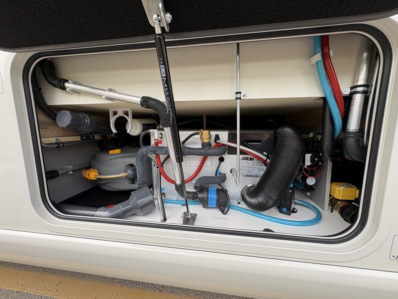 Open exterior storage compartment of a caravan showing plumbing, water hoses, drainage pipes, and an Alde heating system installation.
